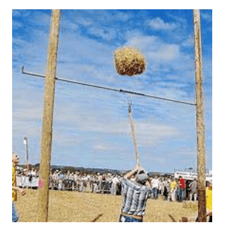Photographie d'une compétition de lancer de gerbe de paille. Une gerbe compacte est en l'air, au-dessus d'une barre horizontale tendue entre deux mâts de bois. Au sol, un lanceur tient une fourche à long manche, entouré de spectateurs.