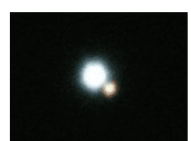 Photographie d'un système d'étoiles doubles sur fond noir : deux taches lumineuses très proches, l'une nettement plus brillante que l'autre.