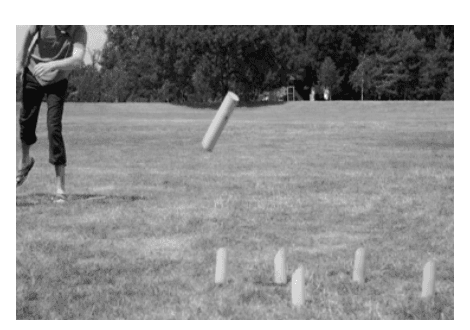 Photographie en noir et blanc d'une pelouse : une personne vient de lancer un bâton de bois, saisi en vol, en direction d'une rangée de quilles cylindriques posées debout sur l'herbe.