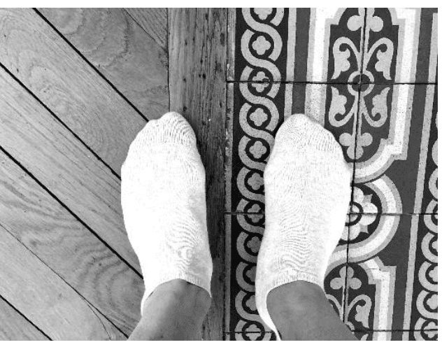 Photographie en noir et blanc de deux pieds chaussés de chaussettes blanches, posés à cheval sur la limite entre un plancher en lames de bois, à gauche, et un carrelage à motifs, à droite.