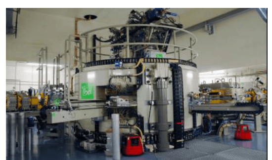 Photographie de l'intérieur d'un hall industriel abritant une machine cylindrique massive entourée de passerelles, de câbles et d'armoires électriques.