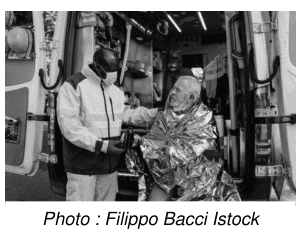 Photographie en noir et blanc d'une intervention de secours : une personne enveloppée dans une couverture de survie métallisée est prise en charge par un secouriste à l'arrière d'un véhicule d'intervention. Photo : Filippo Bacci, Istock.